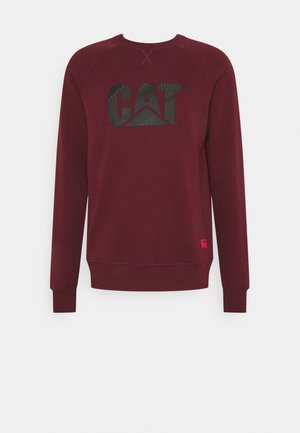Wino czerwona bluza z długimi rękawami i okrągłym dekoltem, z czarnym logo "CAT" na piersi i małą czerwoną prostokątną naszywką blisko dolnego brzegu.