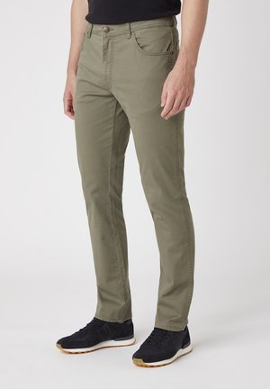 Hombre con pantalones ajustados verde oliva y zapatillas negras, de pie contra un fondo claro y liso.