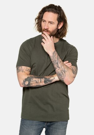 Camiseta de manga corta en verde oliva con cuello redondo, hecha de una tela suave, combinada con jeans azules. Presenta tatuajes visibles en los brazos.