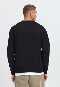 Bărbat cu părul scurt, îmbrăcat cu un hanorac negru peste o cămașă albă și pantaloni bej, întors cu spatele, pe un fundal simplu de culoare deschisă.