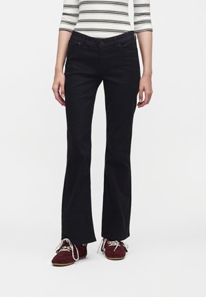 Personne portant un pantalon bootcut noir, un haut à manches longues rayé et des chaussures marron à lacets, debout sur un fond blanc uni.
