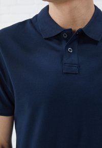 Polo majica tamnoplave boje od teksturiranog materijala, sa klasičnim ovratnikom, dvobotonim kopčanjem i suptilnim detaljima šivanja.