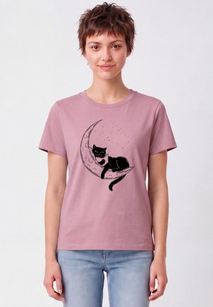 Vrouw met kort bruin haar die een mauve T-shirt draagt met een zwarte kat die ligt te ontspannen op een sikkelmaan-afbeelding, gecombineerd met lichtblauwe spijkerbroek.