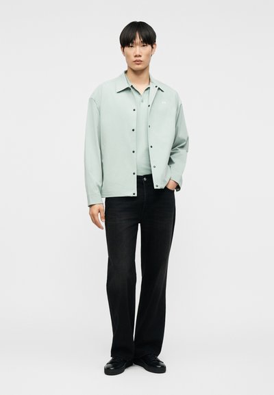 Νεαρός άνδρας που στέκεται, φοράει ένα ανοιχτοπράσινο πουκάμισο με κουμπιά πάνω από ένα αντίστοιχο πόλο, μαύρο παντελόνι και μαύρα παπούτσια, με το ένα χέρι στην τσέπη.