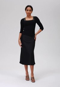 Egy nő áll, fekete négyzetnyakú topot visel gombokkal és fekete midi szoknyát, fekete pántos magassarkúval párosítva, egy egyszínű háttér előtt.