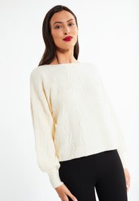 Krém, strukturált pulóver hullámos mintával és ballon ujjakkal, cropped hosszal, fekete leggingsszel párosítva. Puha anyag, laza fazon.