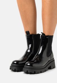 Botas negras brillantes hasta el tobillo con suelas gruesas y de tracción, usadas sobre piernas desnudas, con paneles laterales elásticos y tiradores en la parte posterior.