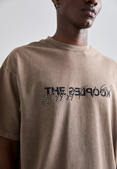 Camiseta de algodón marrón con un diseño gráfico de texto negro decorado con acentos de tachuelas metálicas. Mangas cortas y corte relajado.