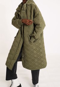 Gequilted olijfgroen lange jas met een capuchon, voorzien van ruitpatronen, ribgebreide manchetten en zijzakken, ritssluiting en een relaxte pasvorm.