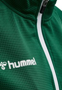 Groene textuur gezoomde jas met een witte rits en een opvallend wit "hummel" logo aan de voorkant.