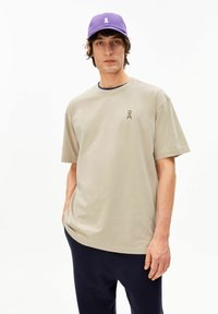 Beige katoenen T-shirt met een losse pasvorm en korte mouwen, met een klein groen logo op de borst. Gecombineerd met een donkerblauwe broek en een paarse pet.