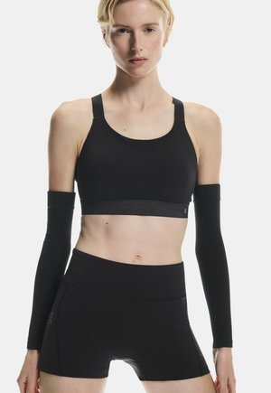 Femme portant un soutien-gorge de sport noir, un short taille haute noir et des manches noires, debout devant un fond clair uni.