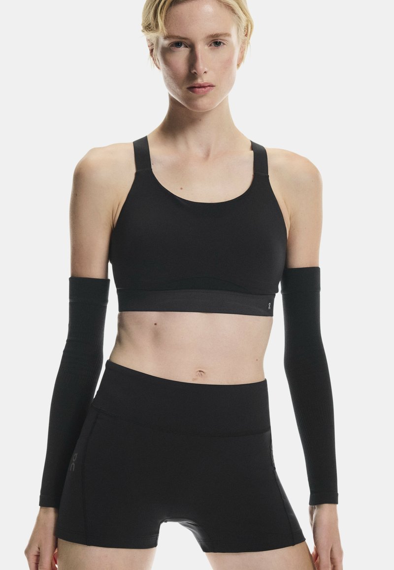 Femme portant un soutien-gorge de sport noir, un short taille haute noir et des manches noires, debout devant un fond clair uni.