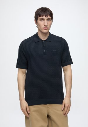 Jeune homme aux cheveux courts et foncés, portant un polo noir texturé à manches courtes et un pantalon beige, debout devant un fond blanc.