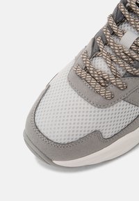 Pointe de chaussure de sport grise et beige avec tissu en mesh, superpositions en suède et lacets à motifs sur fond blanc.