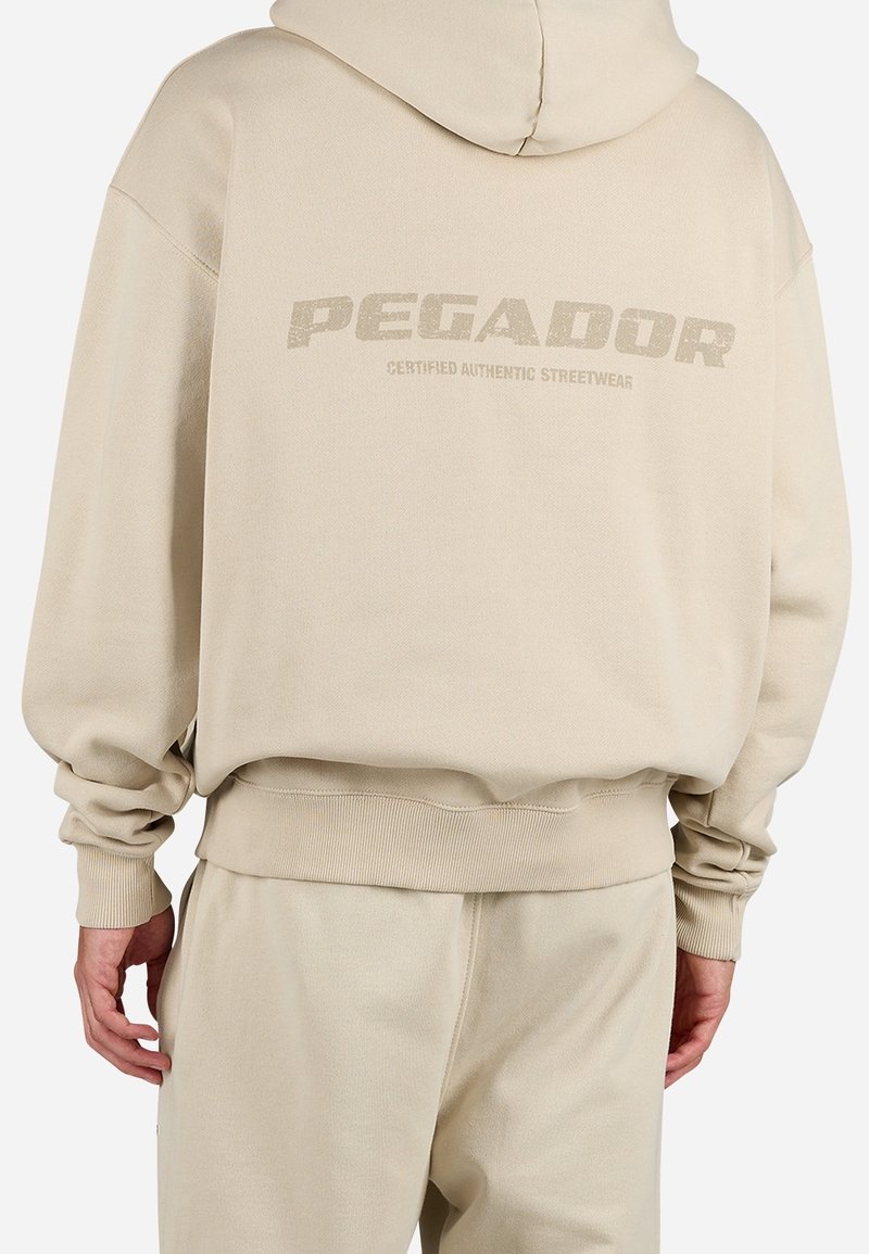 Sweat à capuche beige et pantalon assorti portés par une personne, avec "PEGADOR CERTIFIED AUTHENTIC STREETWEAR" imprimé dans le dos.