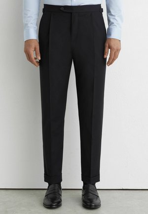 Mężczyzna ubrany w dopasowane czarne spodnie garniturowe z mankietami, jasnoniebieską koszulę w paski oraz czarne skórzane mokasyny, stojący na tle białej ściany.