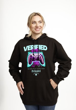 Femme souriante portant un sweat à capuche noir avec un graphisme néon d'une manette de jeu violette et le texte « JOUEUSE VÉRIFIÉE, Pro Joueur Niveau 55, Préchargé. »