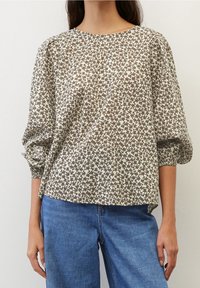 Blusa a maniche lunghe di colore verde chiaro con motivo floreale marrone. Vestibilità comoda con maniche arricciate e scollatura rotonda. Indossata con jeans blu.