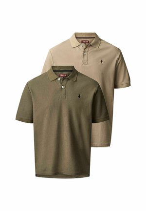 Homme aux longs cheveux et tatouages porte un polo vert olive, un jean noir et des bottes marron, debout devant un fond uni clair.
