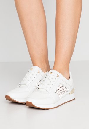 White lace-up sneakers with mesh side panels, gold accents, and brown soles worn on bare legs against a neutral background.