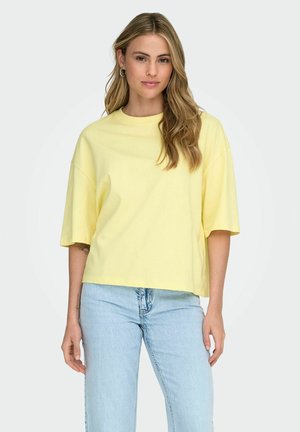 Junge Frau mit langen, welligen Haaren, die ein locker sitzendes, hellgelbes T-Shirt und hellblaue Jeans trägt und entspannt steht.