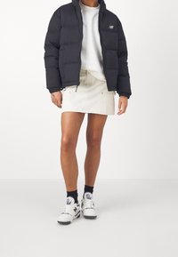 Osoba ubrana w czarną kurtkę puchową, białą bluzę, beżową mini spódnicę, czarne skarpetki i białe sneakersy stoi na białym tle.