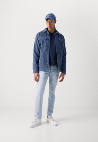 Veste en velours côtelé bleu avec deux poches poitrine, un pull marine à col rond, un jean bleu clair et des baskets blanches, le tout agrémenté d'une casquette bleue.