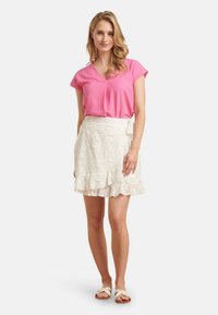 Rosa kurzärmliges Oberteil, kombiniert mit einem weißen Rüschensrock mit floralen Mustern. Trägt helle Sandalen.