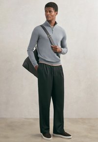 Mężczyzna ubrany w jasnoszary sweter z zamkiem przy szyi, czarne szerokie spodnie, brązowe trampki, niosący czarną torbę na ramię, stojący na jednolitym tle.