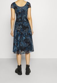 Robe bleue à motifs cachemire, avec de courtes manches en maille, un corsage ajusté, une jupe évasée et des bottines noires. Tissu lisse avec un léger reflet.