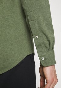 Zielona bluzka z długim rękawem i białymi guzikami na lewym ramieniu, zestawiona z czarnymi spodniami na jednolitym jasnym tle.