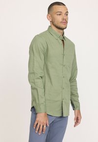 Chemise boutonnée vert clair en tissu texturé, dotée d'une poche poitrine et d'un col boutonné, portée avec un pantalon bleu.