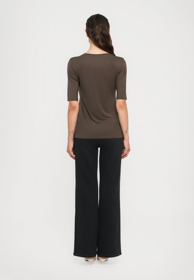 Donna con lunghi capelli scuri rivolta di spalle, che indossa una maglietta marrone a maniche elbow, pantaloni neri a gamba larga e tacchi beige, in piedi contro uno sfondo bianco.