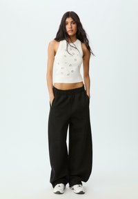 Ung kvinna med långt mörkt hår, iförd en ärmlös vit crop top med metallöglor, svarta vida byxor och vita sneakers.