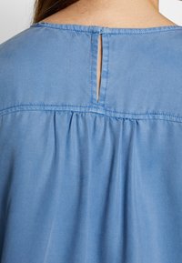 Lys blå bluse med nøglehul detalje på ryggen, samlede plisseringer ved halsen og en glat, let stofstruktur.