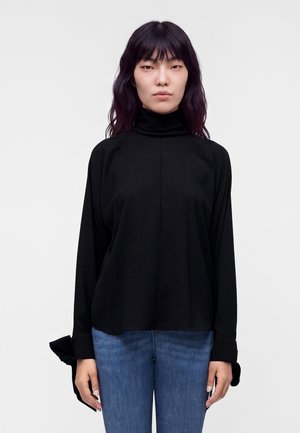 Voksen kvinde med sort højhalset langærmet top og blå jeans stående mod en ensfarvet lys baggrund.