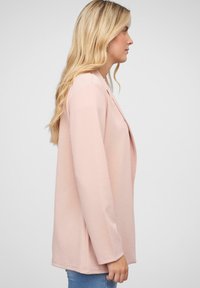 Lys pink, langærmet jakke med en afslappet pasform. Glat tekstur, åben frontdesign og rundet kant. Set fra siden.