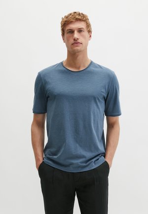 Jonge man met kort krullend blond haar, draagt een blauw T-shirt met korte mouwen en een zwarte broek, staat met handen in de zakken.