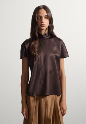 Femme aux longs cheveux foncés portant un haut en satin marron foncé à manches courtes et une jupe plissée taille haute couleur beige sur un fond uni.