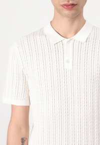 Hvid strikket polo-shirt med vertikal ribstruktur, korte ærmer og en klassisk krave med tre knapper.