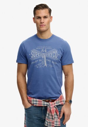 Homme portant un t-shirt bleu Superdry avec un motif de phare, une chemise à carreaux nouée autour de la taille, un jean bleu et une montre-bracelet au poignet gauche.