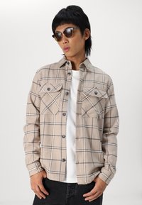 Beige geruite flanellen shirt met contrasterende zwarte lijnen, twee borstzakken, knoopsluiting aan de voorkant en een zachte textuur. Draagt over een wit T-shirt.