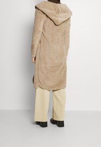 Beżowy płaszcz z sztucznego futra o długim kroju, z dużym kapturem, teksturowaną powierzchnią i bocznymi rozcięciami. Połączony z kremowymi spodniami w szerokim kroju i ciemnymi, platformowymi butami.