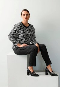 Zwart-witte blouse met patroon en lange mouwen, gecombineerd met zwarte slim fit broek en glanzende zwarte hoge hakken met kettingdetail.