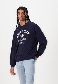 Sudadera azul marino con el texto "NEW YORK GAP ATHLETIC DEPT.", puños y dobladillo acanalados, combinada con jeans de color azul claro.