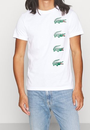 Biała bawełniana koszulka z okrągłym dekoltem, z czterema małymi zielonymi grafikami krokodyli po lewej stronie. Zestawiona z jasnoniebieskimi dżinsami.