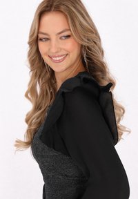 Top negru cu volane din material transparent, combinat cu un corset argintiu strălucitor. Părul ondulat și cerceii lungi completează look-ul.