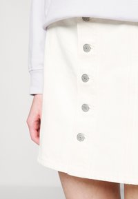 Jupe en denim blanc avec des boutons-pression argentés sur le devant, un ourlet légèrement évasé et une texture lisse. Associée à un haut violet clair.