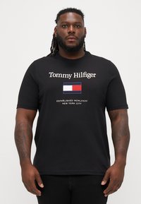 Mustmustäheline T-särk, millel on "Tommy Hilfiger" logo ja silindrikujuline lipugraafika sinises, valges ja punases. Lühikesed varrukad ja ümmargune kaelus.